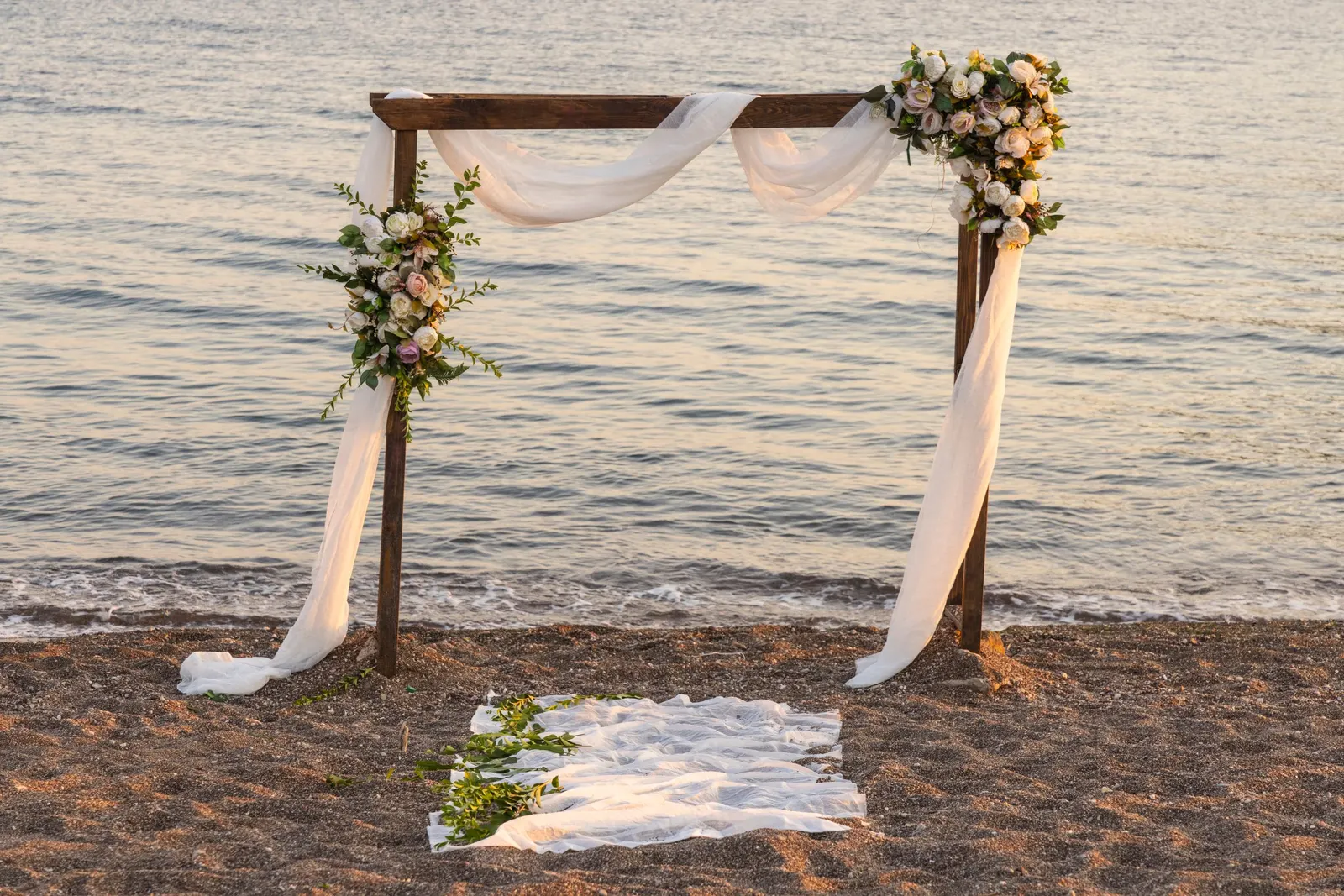 Wooden wedding arch with white drapes and flowers on the sand at the water's edge
