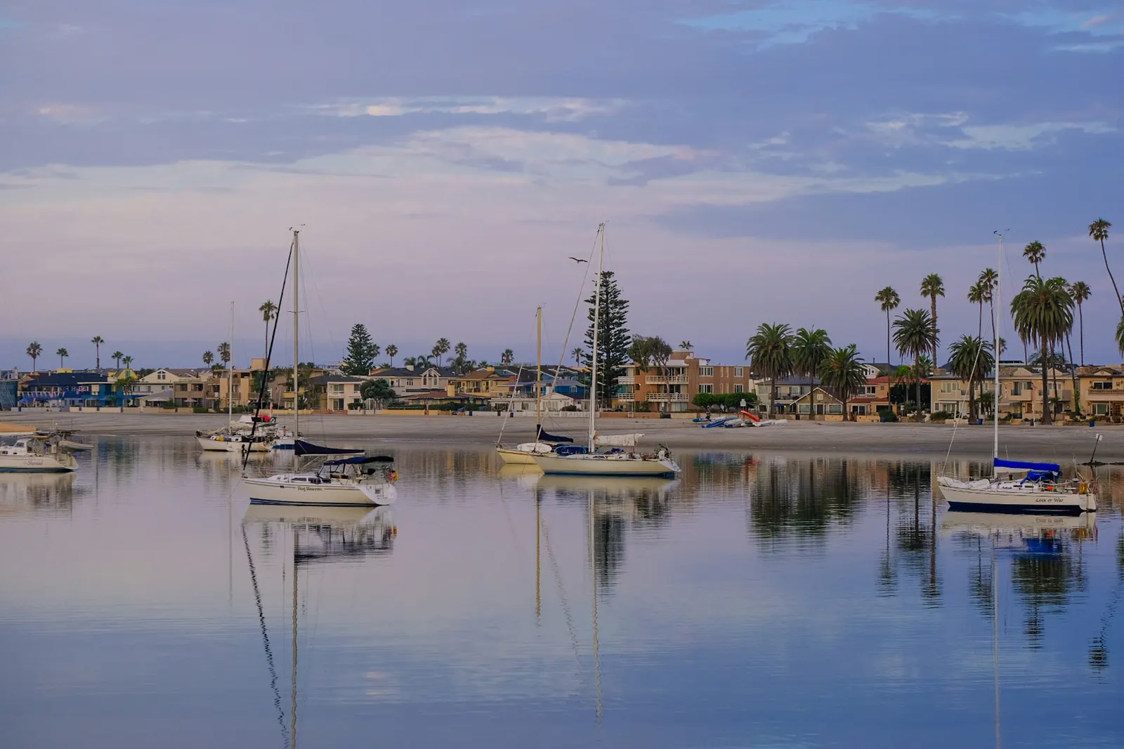 Sailboats moored in calm Mission Bay at dusk, with beachfront homes and palms