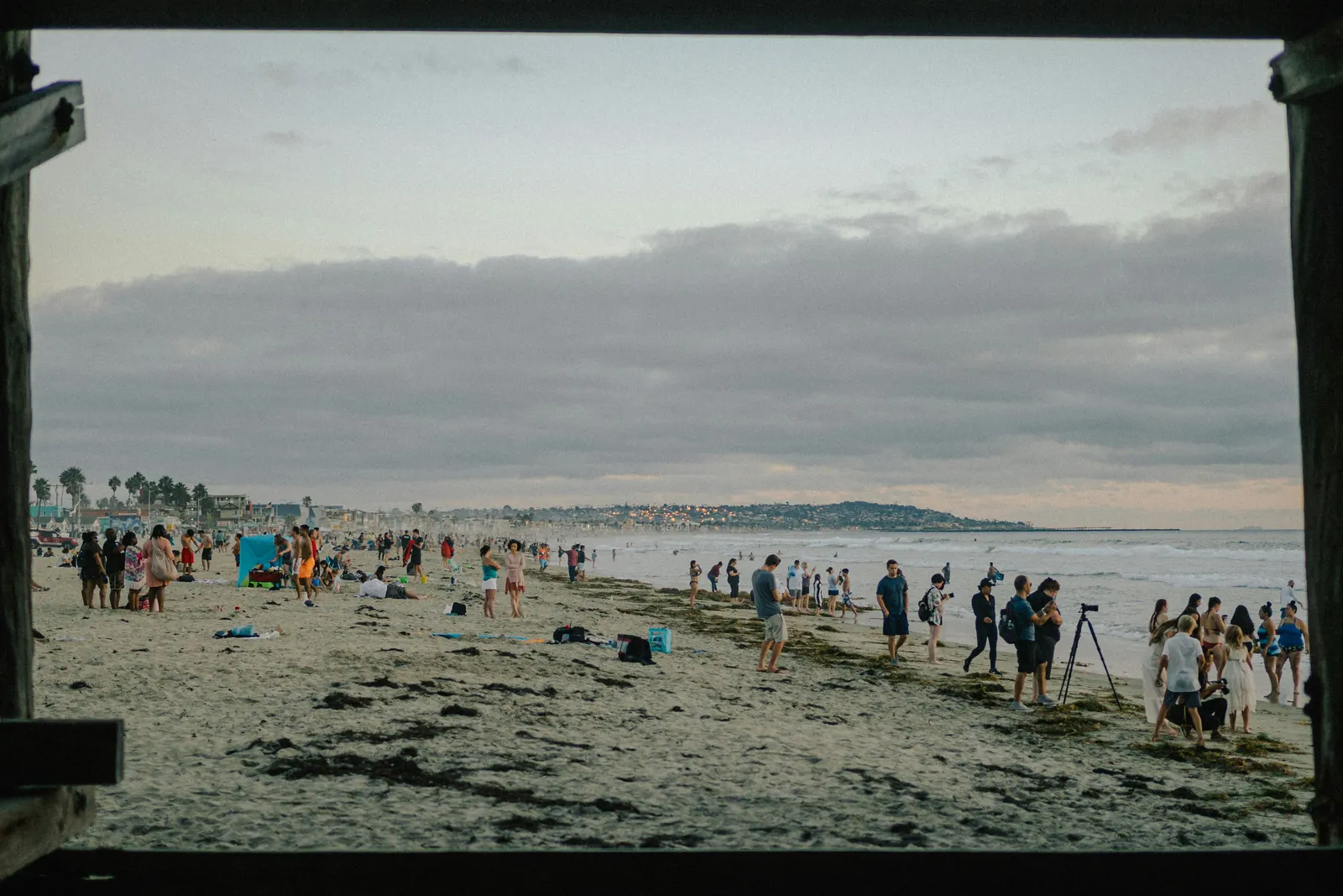Groups of people gathered on Pacific Beach at dusk, seen from under the pier